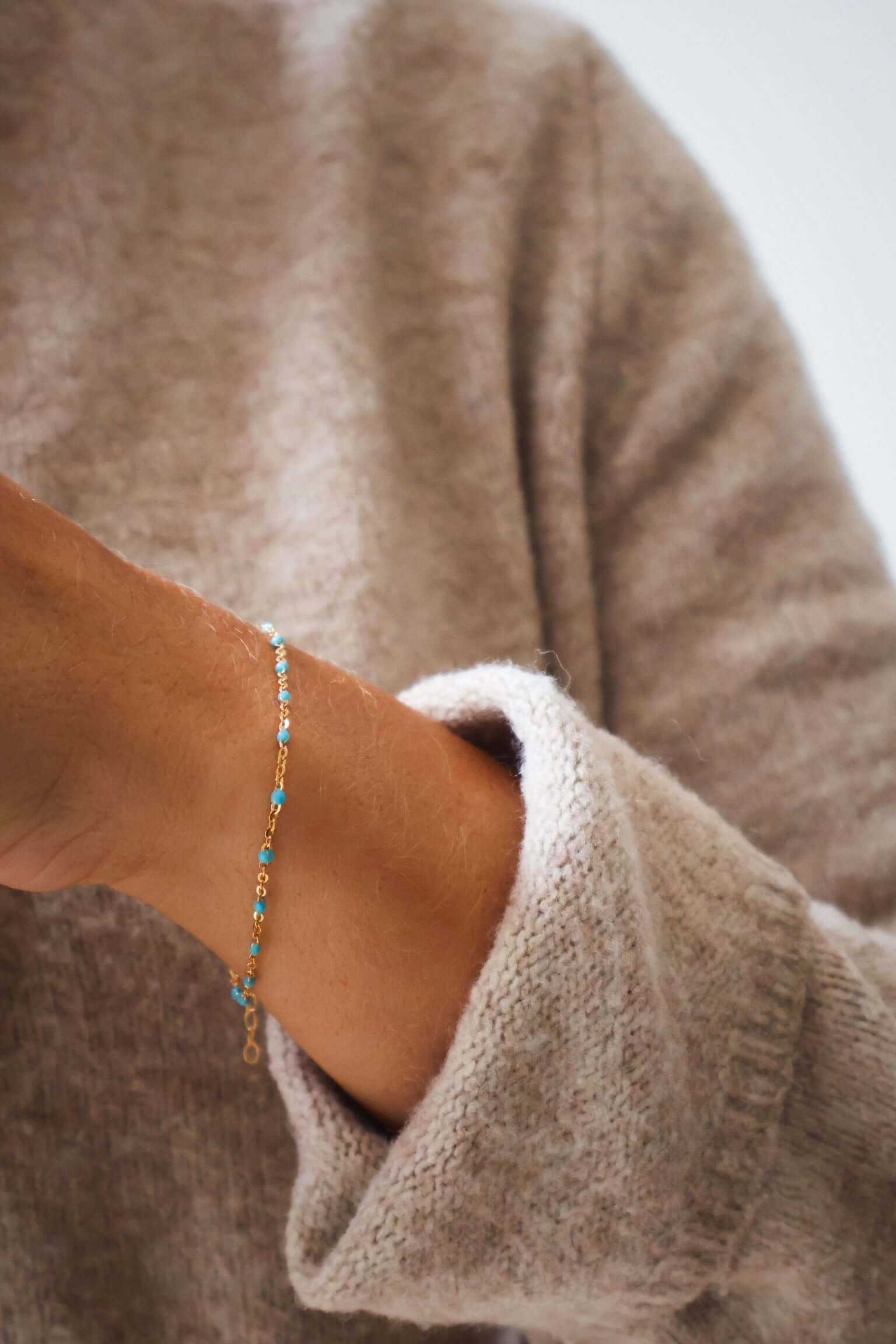 Close-up of a hand wearing a 14k gold turquoise beaded bracelet with a cozy sweater, showcasing ocean-inspired color.