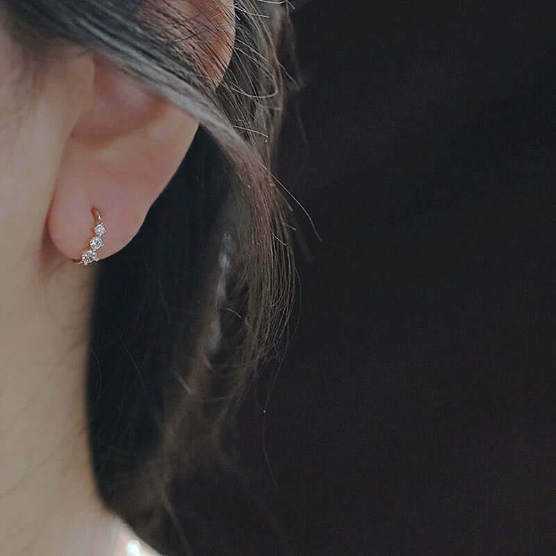Tiny gold huggie half hoop earrings with three cubic zirconia stones, showcased on an ear.