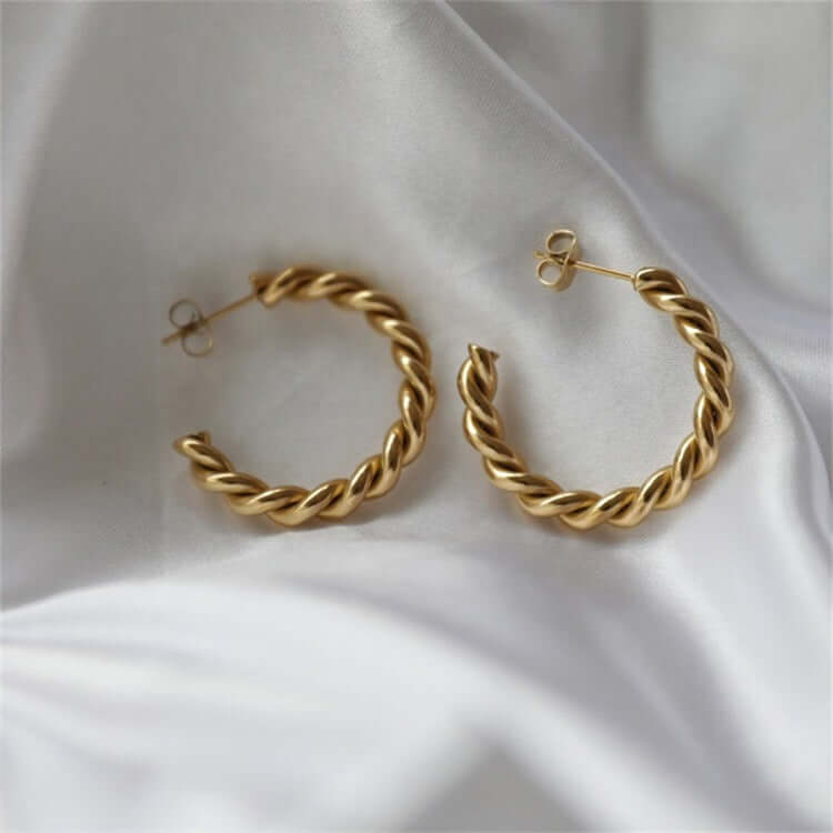 Twisted Gold Hoop Earrings on a soft gray fabric background, showcasing their elegant design and shiny finish.