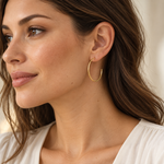 A close-up of a woman wearing a delicate, thin gold hoop earring on her ear, shown in profile against a soft, light beige background.