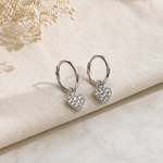 Heart-shaped silver earrings on a soft beige fabric background