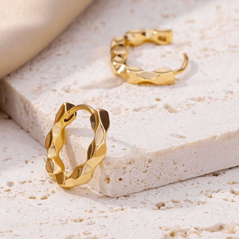Gold hammered effect huggie hoop earrings displayed on a beige surface, showcasing their elegant design.
