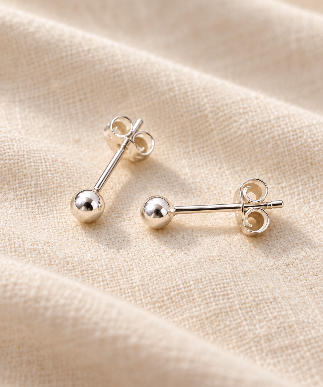 A pair of silver stud earrings with spherical ball studs displayed on a textured beige fabric background.