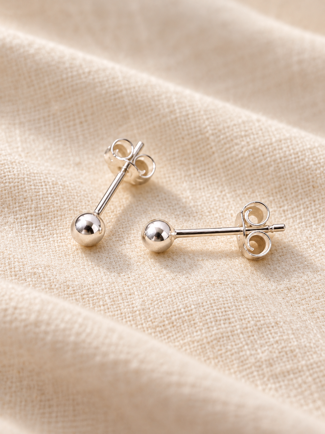 A pair of silver stud earrings with spherical ball studs displayed on a textured beige fabric background.