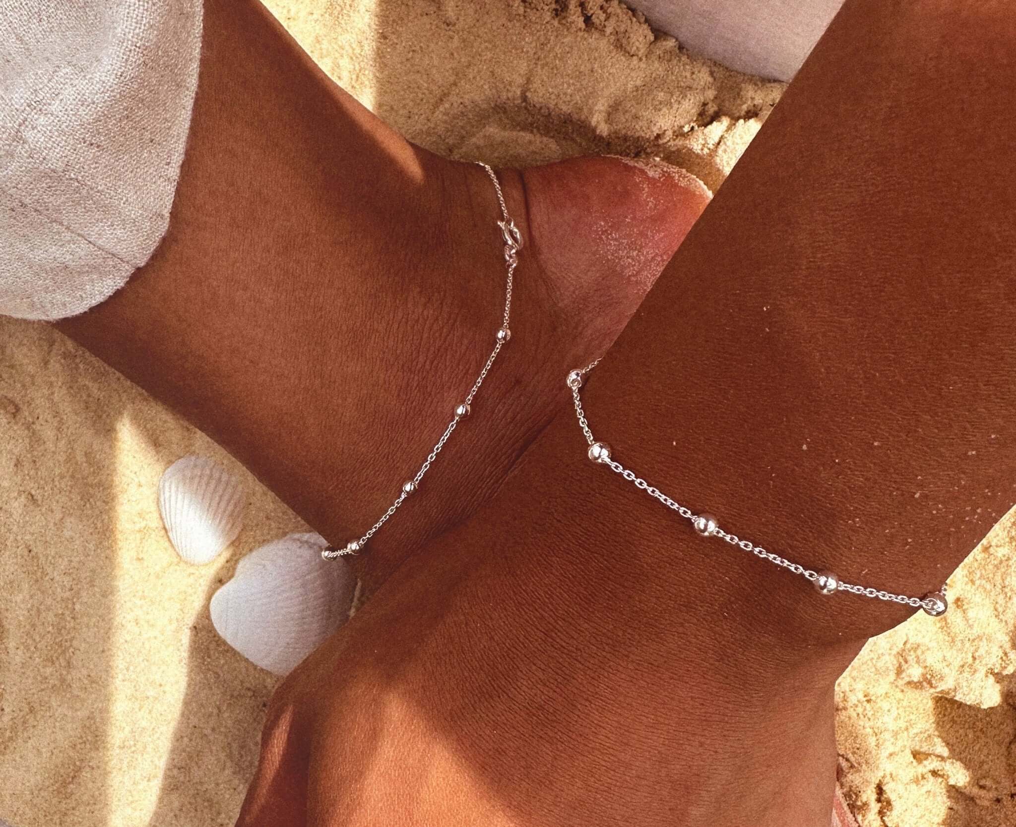 Sterling Silver Chunky Satellite Anklet worn on tanned ankles against sandy beach background with seashells.